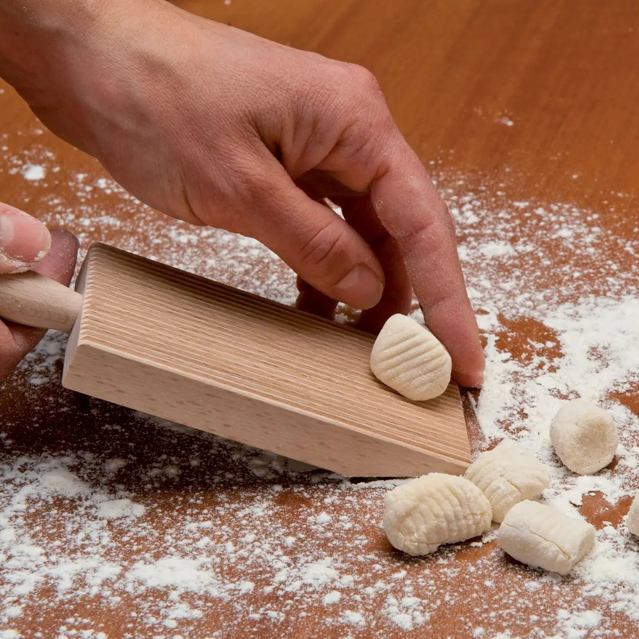 Hand rolling a piece of gnocchi dough down the ridged paddle, held at an angle on a floured wooden worktop