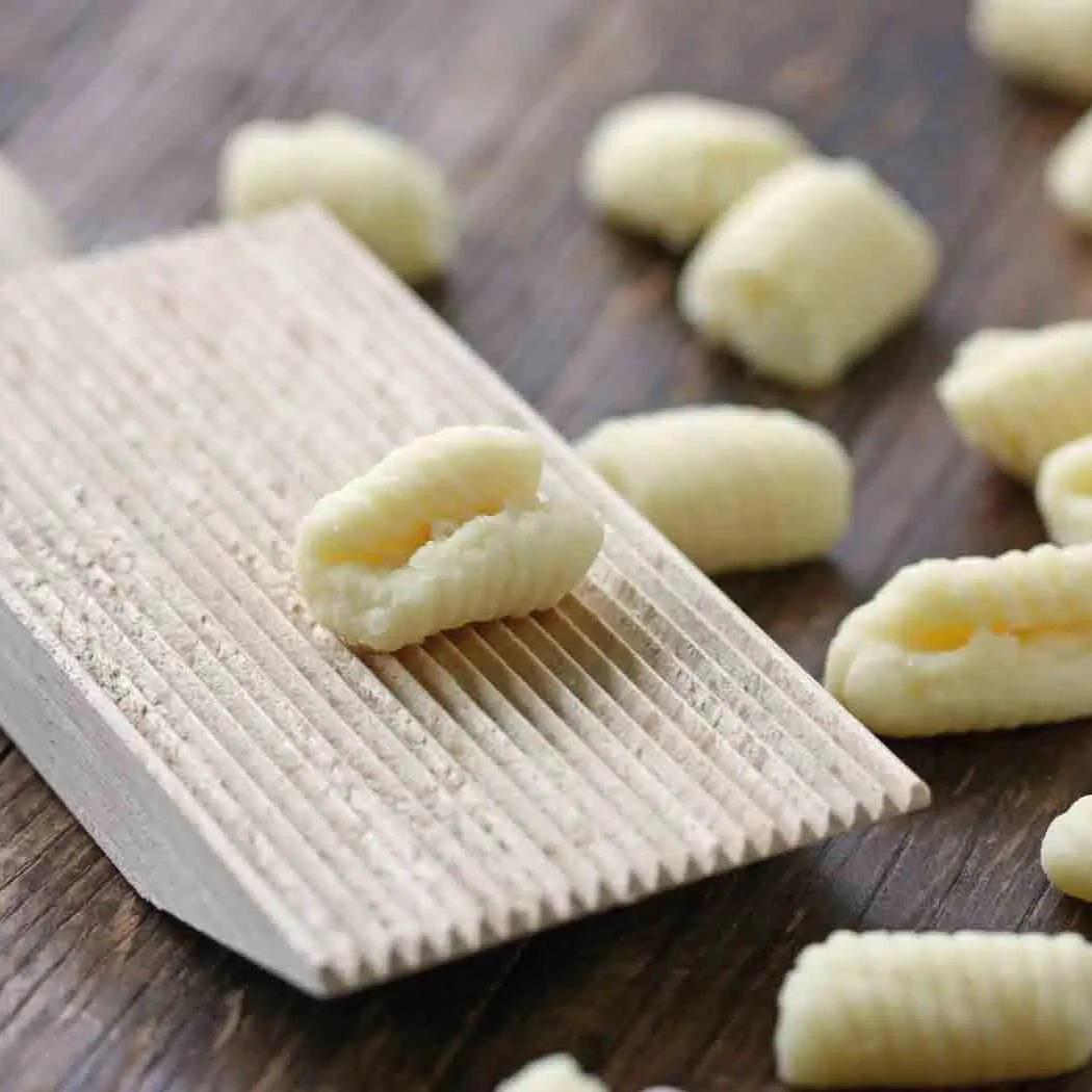 Ridged wooden gnocchi paddle with a ridged piece of gnocchi on its surface and more gnocchi scattered on a dark wooden worktop
