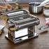 Pasta maker on a wooden table with fresh pasta and kitchen items in the background