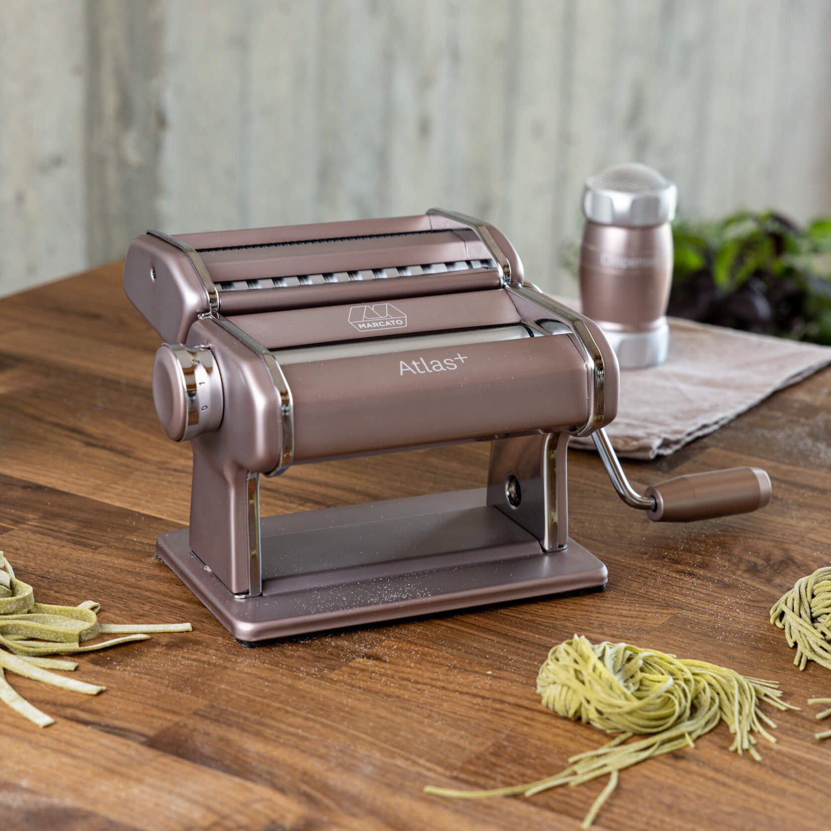 Powder pink Atlas+ pasta machine on a wooden worktop, in front of fresh pasta