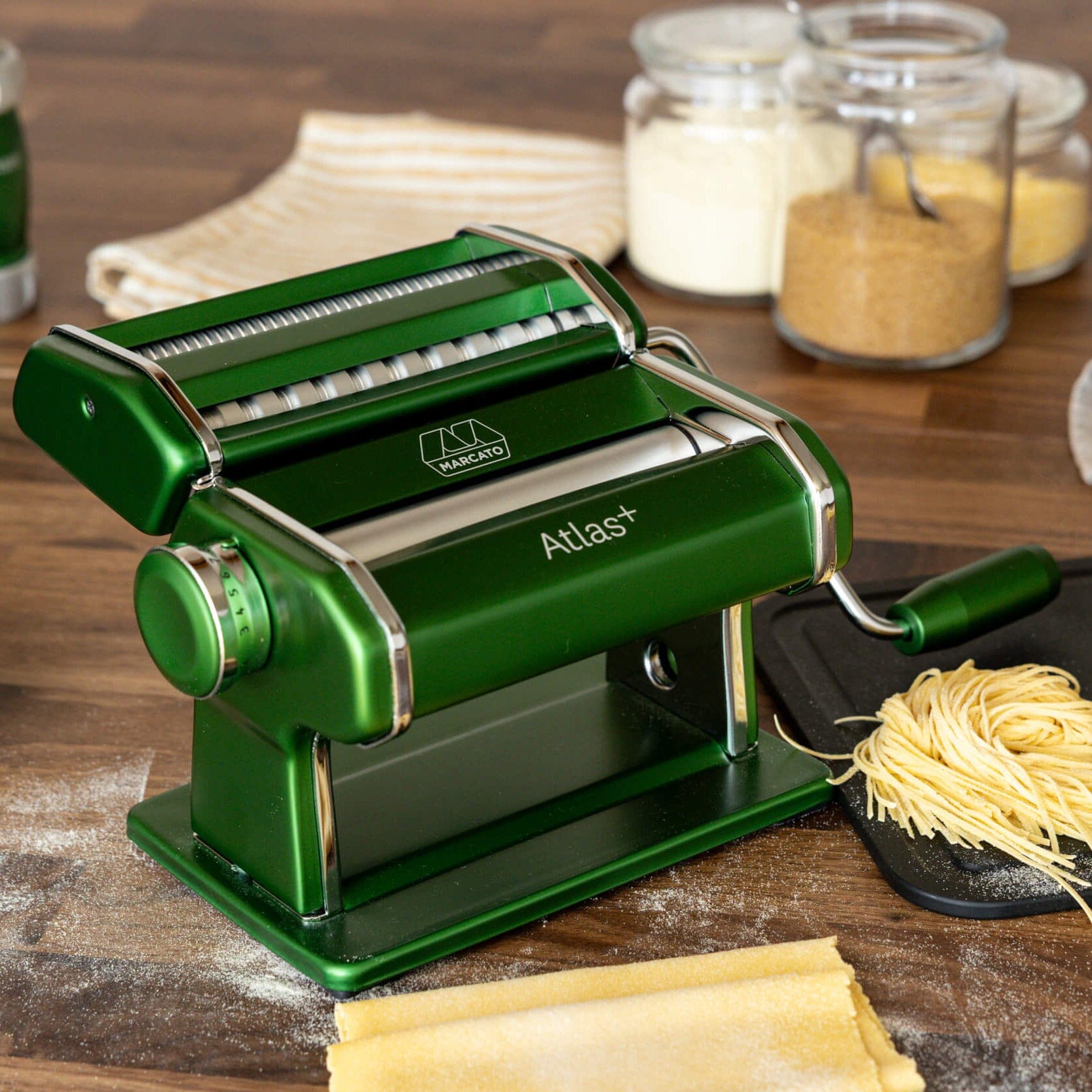 Green pasta maker on a wooden surface with fresh pasta and ingredients.