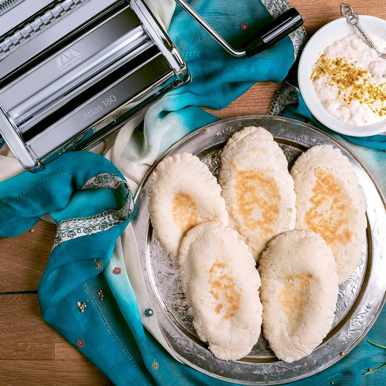 Marcato Atlas 180 pasta maker on a blue textile, next to homemade pitta breads and a dip.