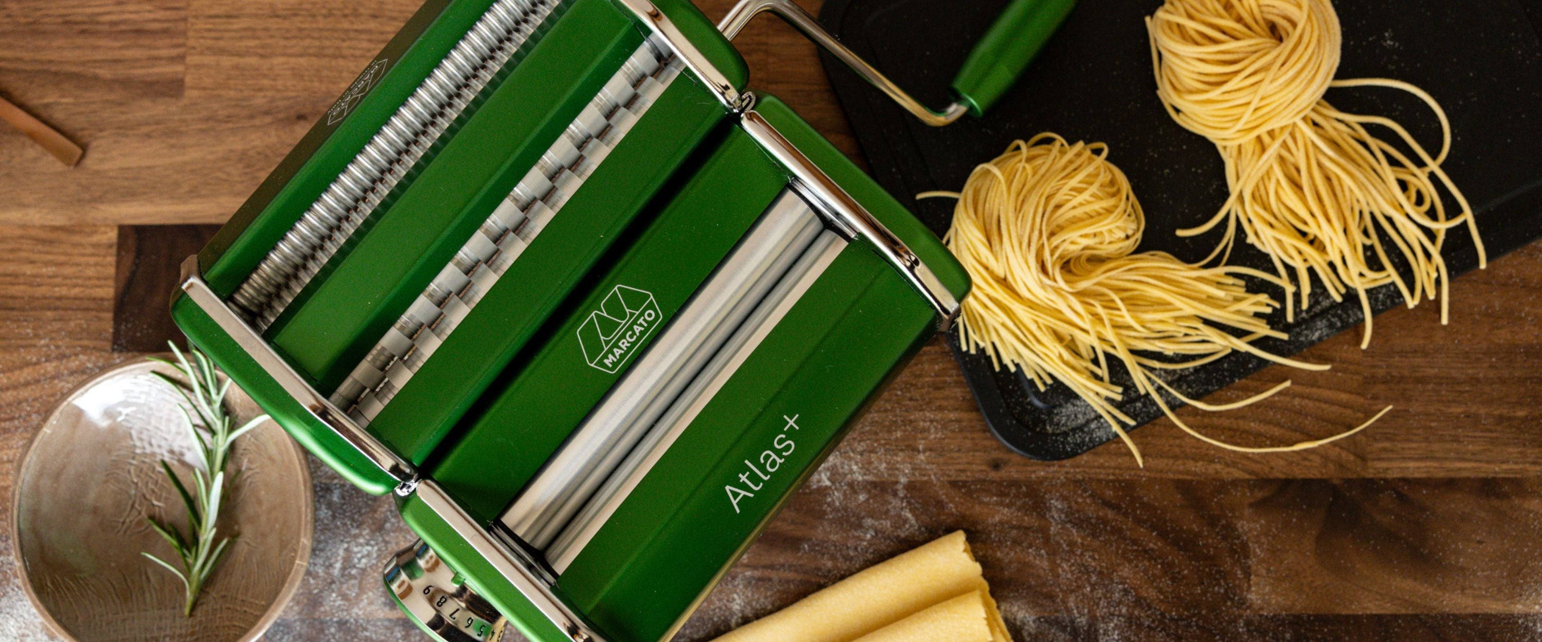 Green Marcato Atlas+ pasta machine on a wooden worktop with fresh tagliatelle, rolled pasta sheets and a sprig of rosemary.