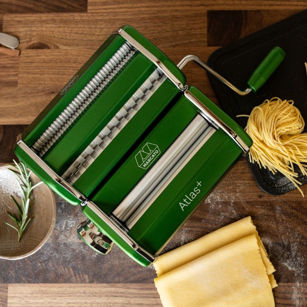 Green Marcato Atlas+ pasta machine on a wooden worktop with fresh tagliatelle, rolled pasta sheets and a sprig of rosemary.