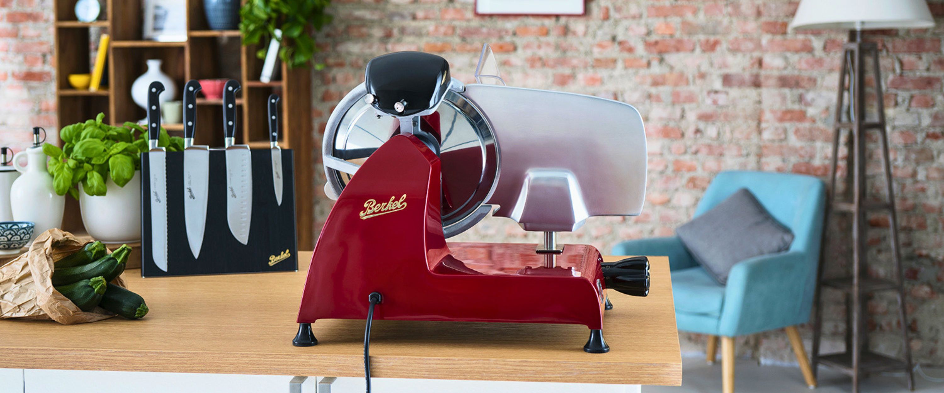 Red Berkel flywheel slicer on a kitchen counter with a Berkel knife block, courgettes and fresh basil in the background.