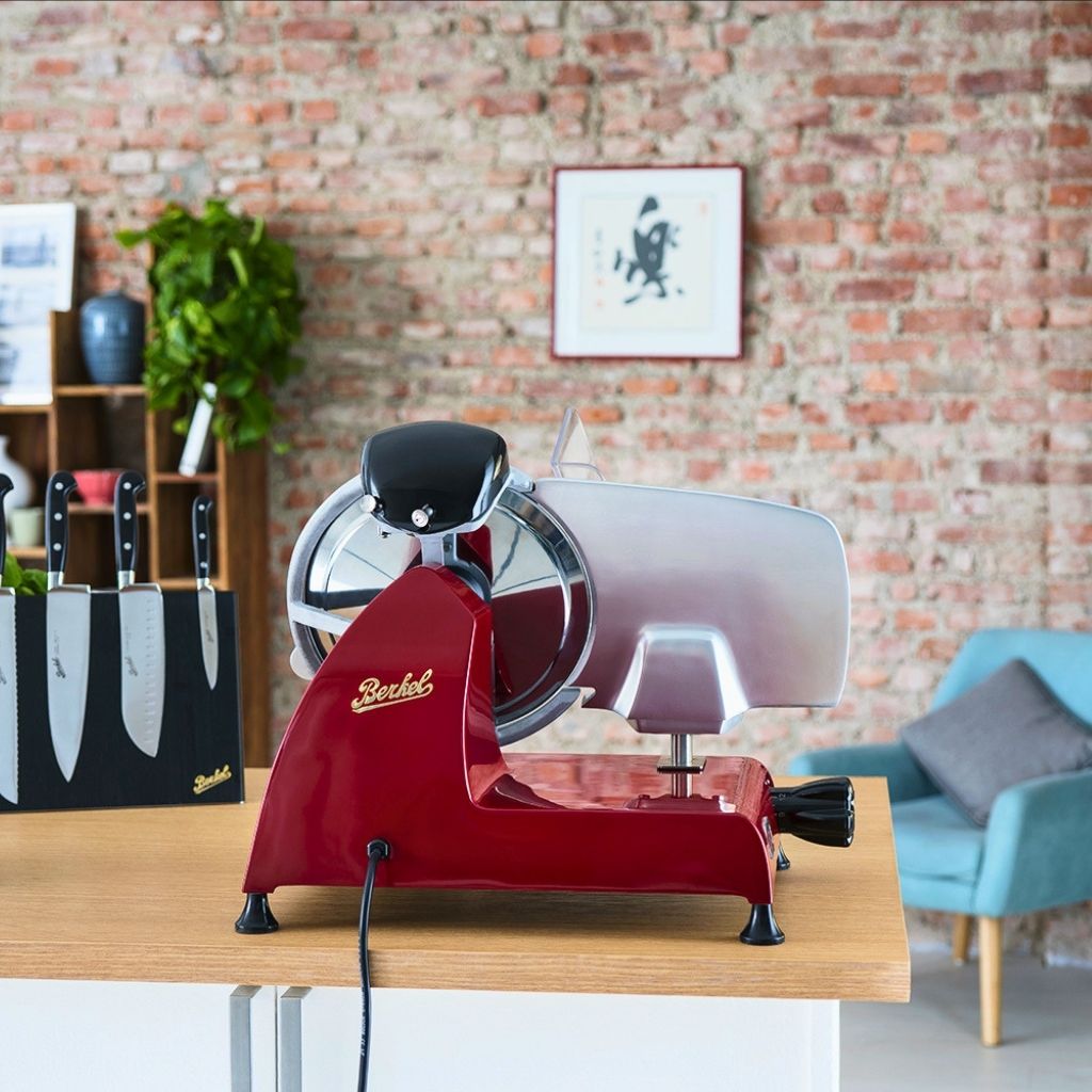 Red Berkel flywheel slicer on a kitchen counter with a Berkel knife block, courgettes and fresh basil in the background.