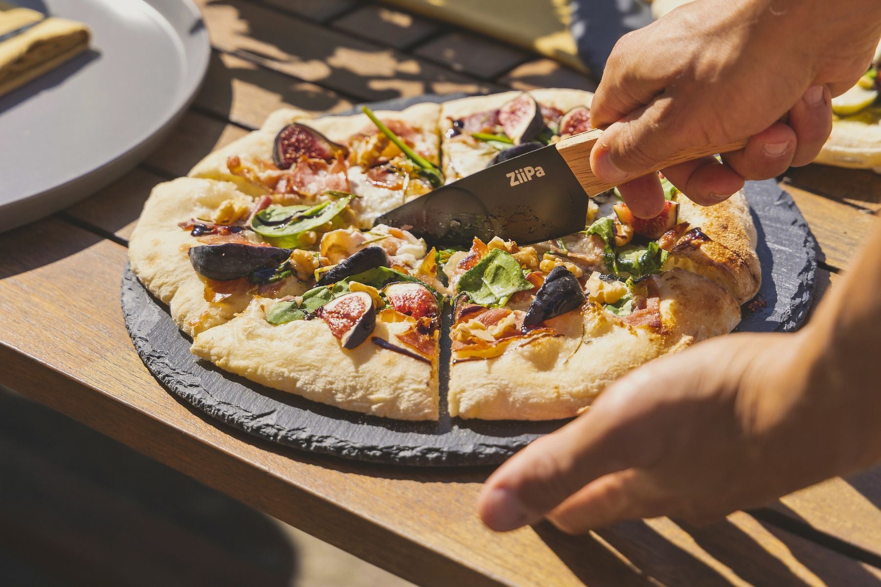 ZiiPa Aquina pizza cutting knife being used on a cooked pizza on a slate serving board on a wooden table top