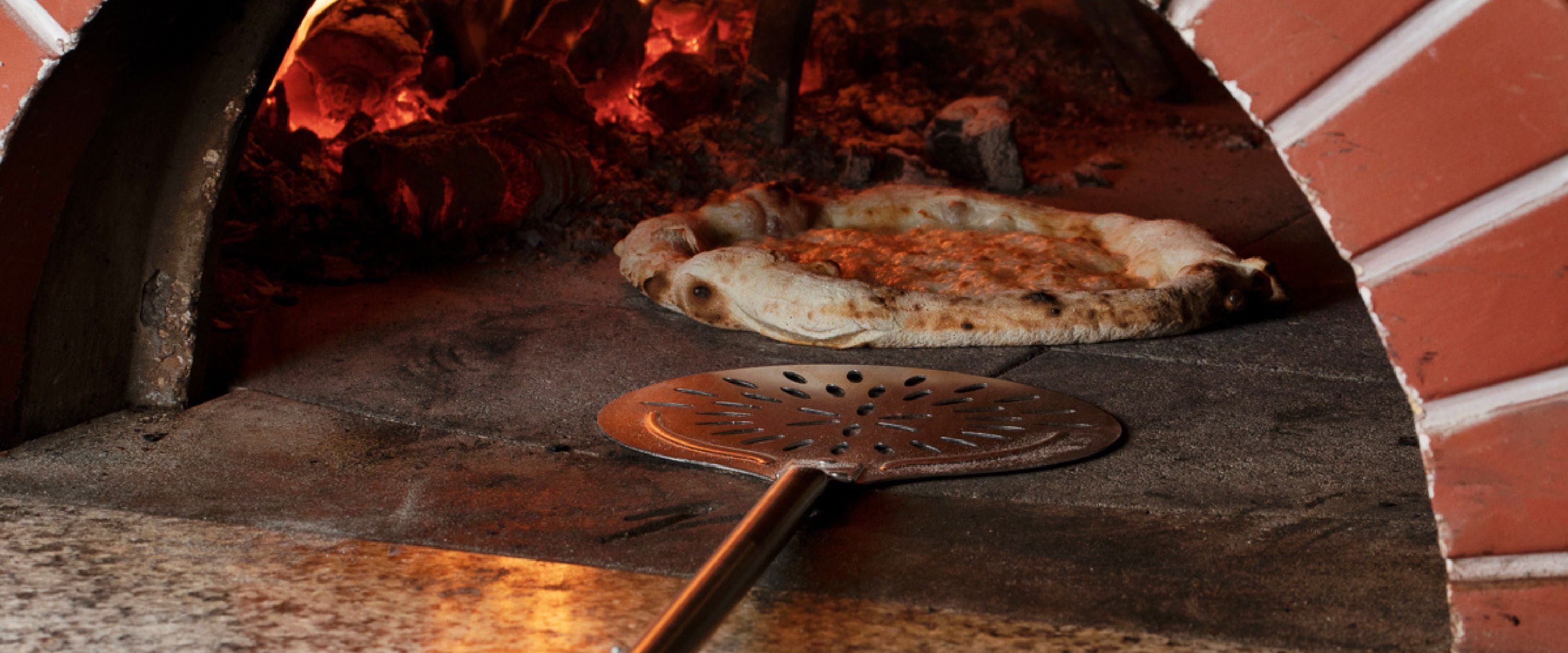 Cerutti Inox pizza turner with a perforated blade, resting in the mouth of a wood-fired oven beside a cooked pizza.