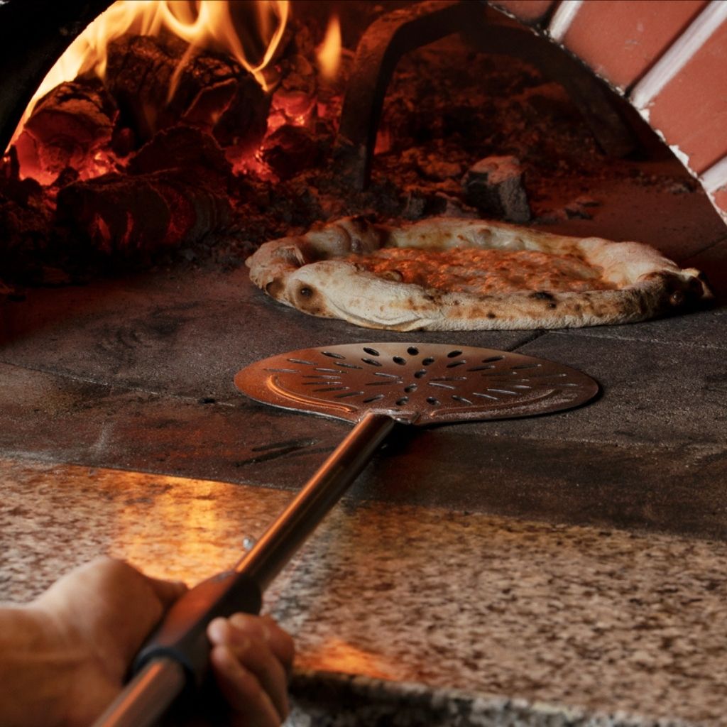 Cerutti Inox pizza turner with a perforated blade, resting in the mouth of a wood-fired oven beside a cooked pizza.