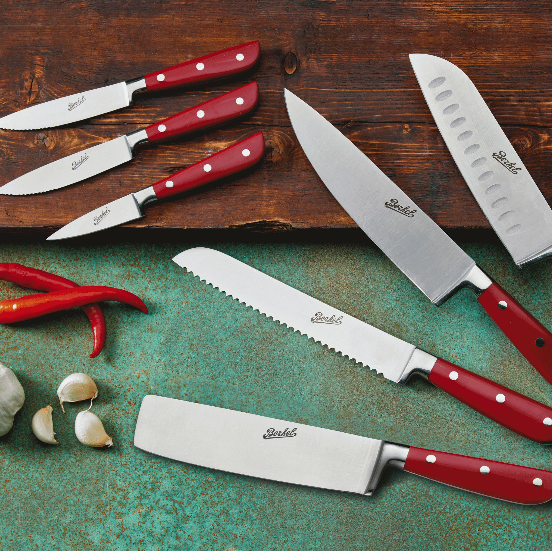 Collection of Berkel Classic Line kitchen knives on a wooden chopping board and blue work surface next to garlic cloves and chillies.