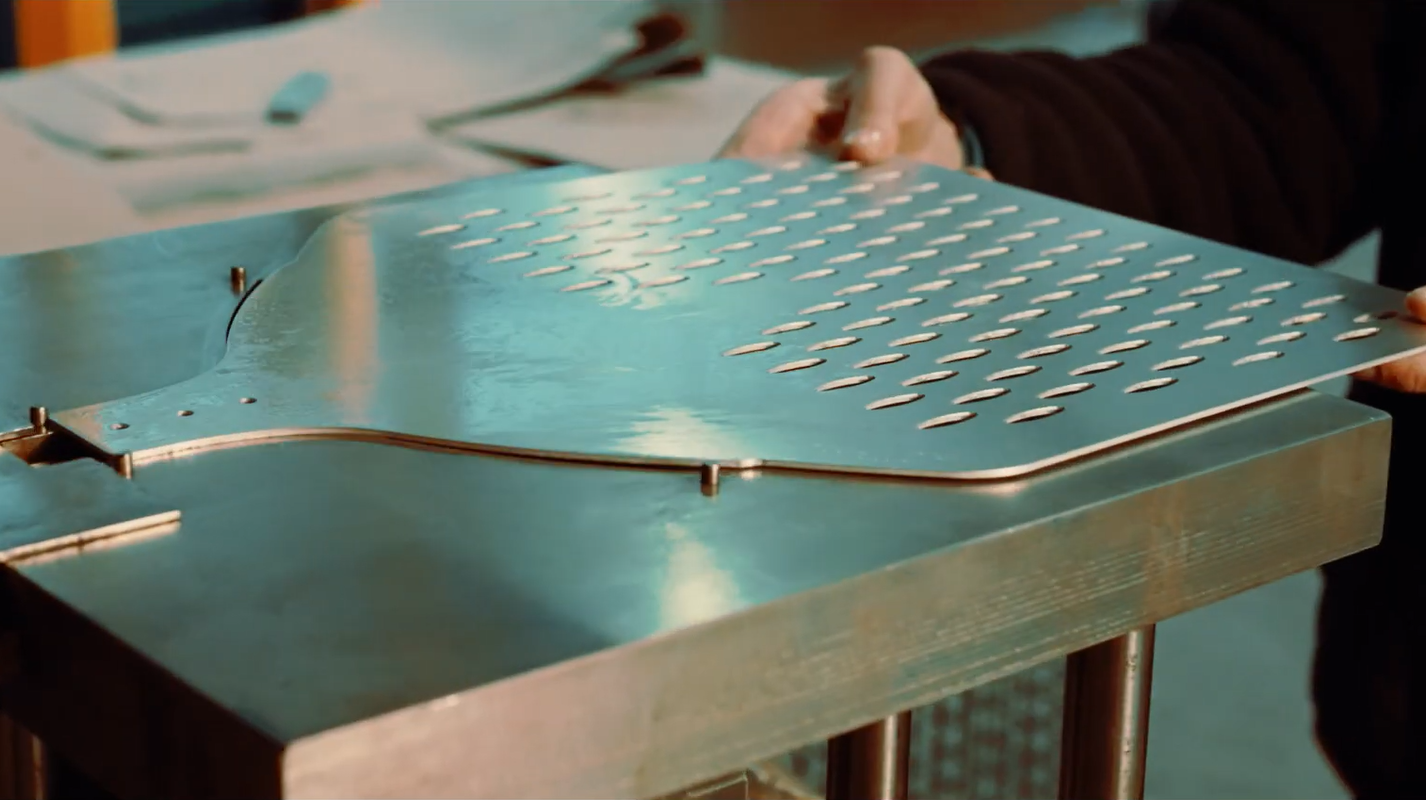 A person holds a large perforated pizza peel sheet over a metal workbench in an industrial or workshop setting.