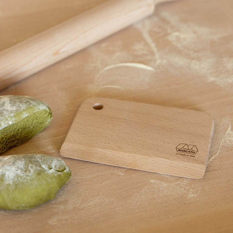 Wooden dough scraper with Marcato logo next to a rolling pin and fresh pasta on a wooden bench
