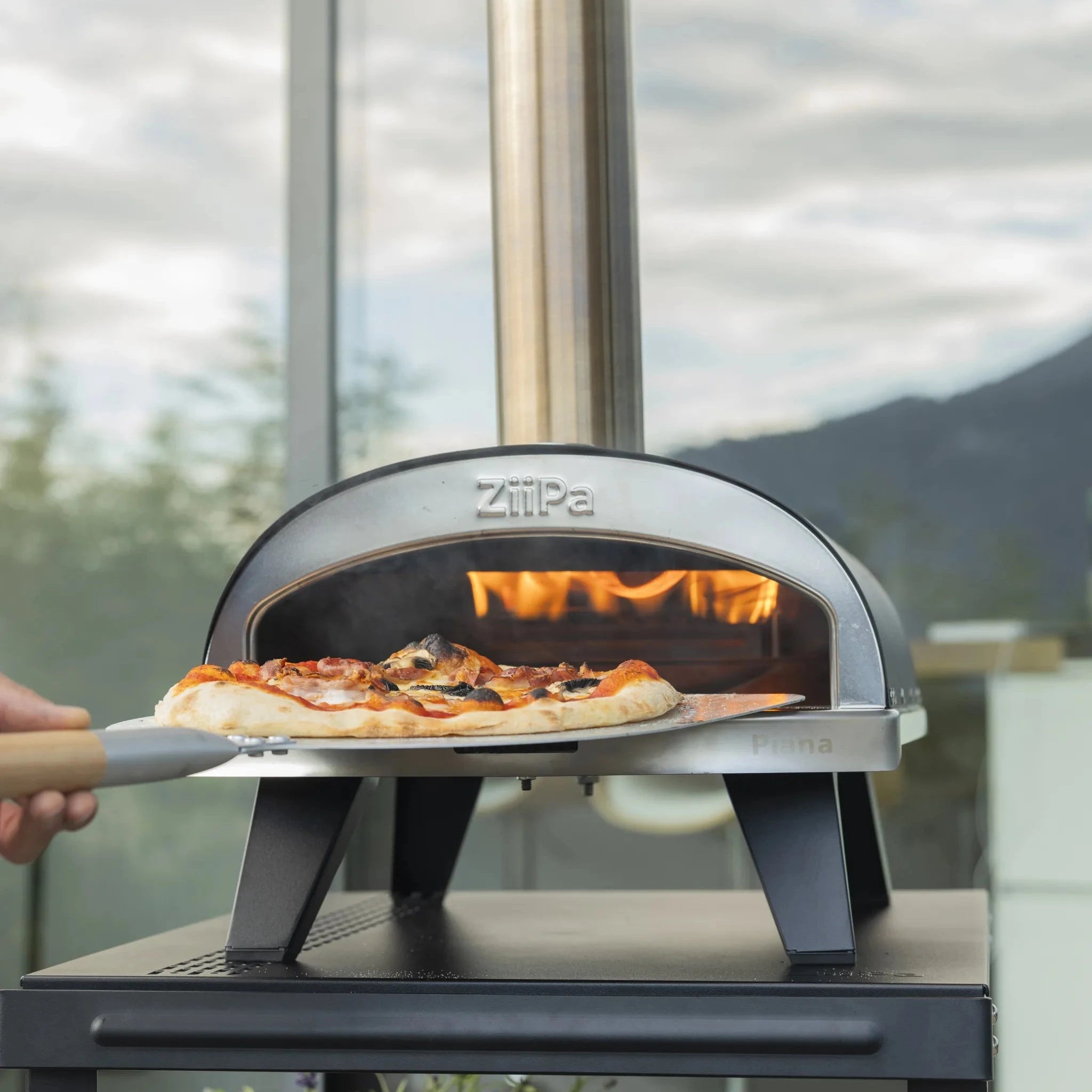 ZiiPa Piana wood fired pizza oven with a pizza being inserted, set against a blurred background.