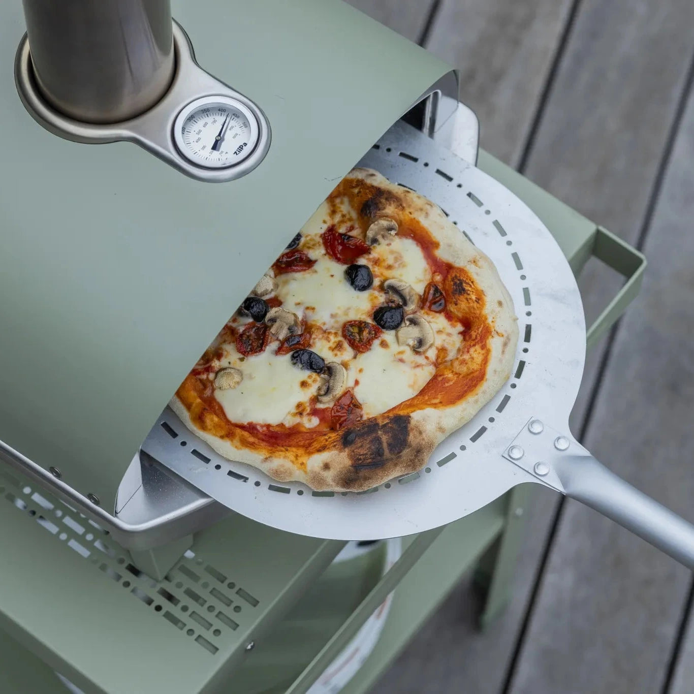 Eucalyptus ZiiPa Piana wood-fired pizza oven outdoors on a wooden decking with a pizza being removed after cooking.