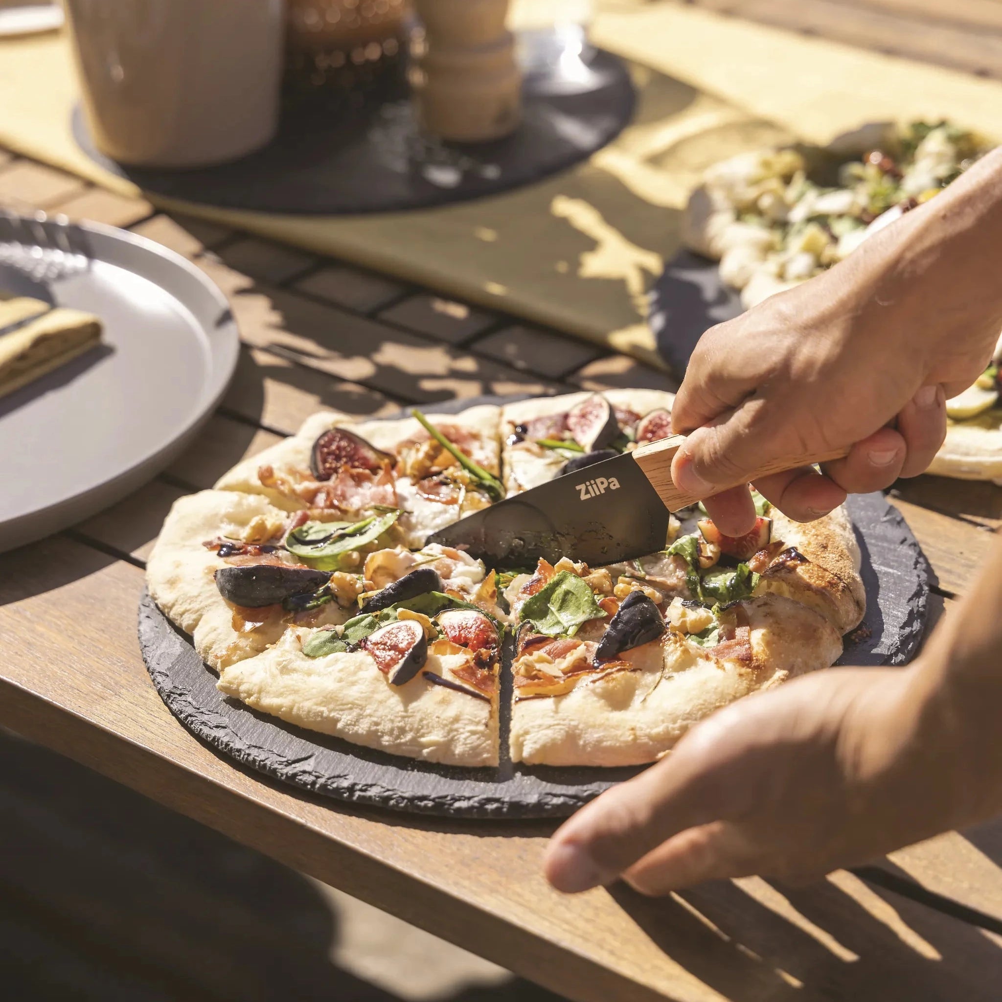 ZiiPa Aquina pizza cutting knife being used on a cooked pizza on a slate serving board on a wooden table top