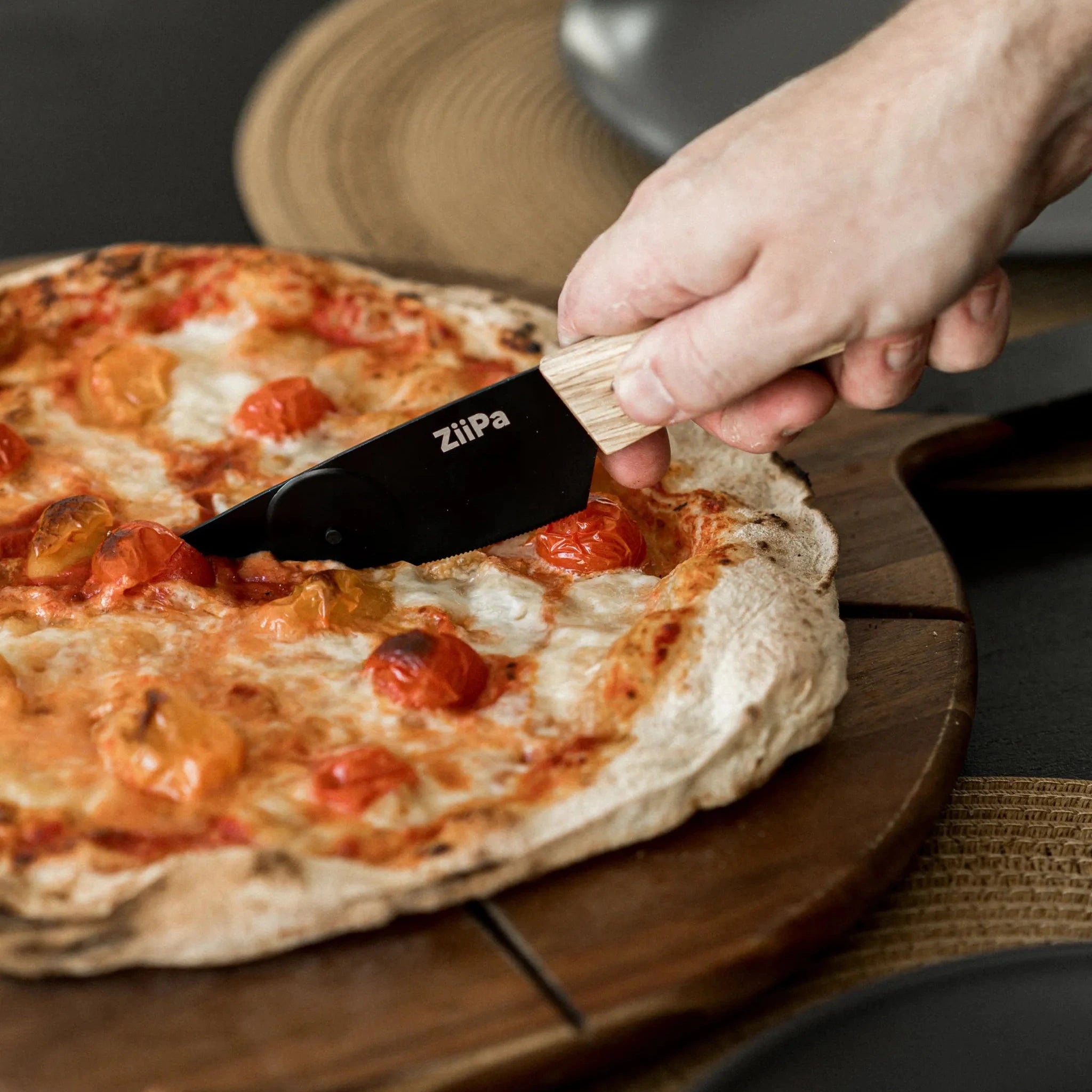 Close-up of a ZiiPa Aquina pizza cutting knife being used on a cooked pizza on a wooden serving board