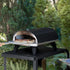 ZiiPa Piana wood-fired pizza oven on a vallone trolley, both in Charcoal colour, in a garden setting with a pizza being removed from the oven