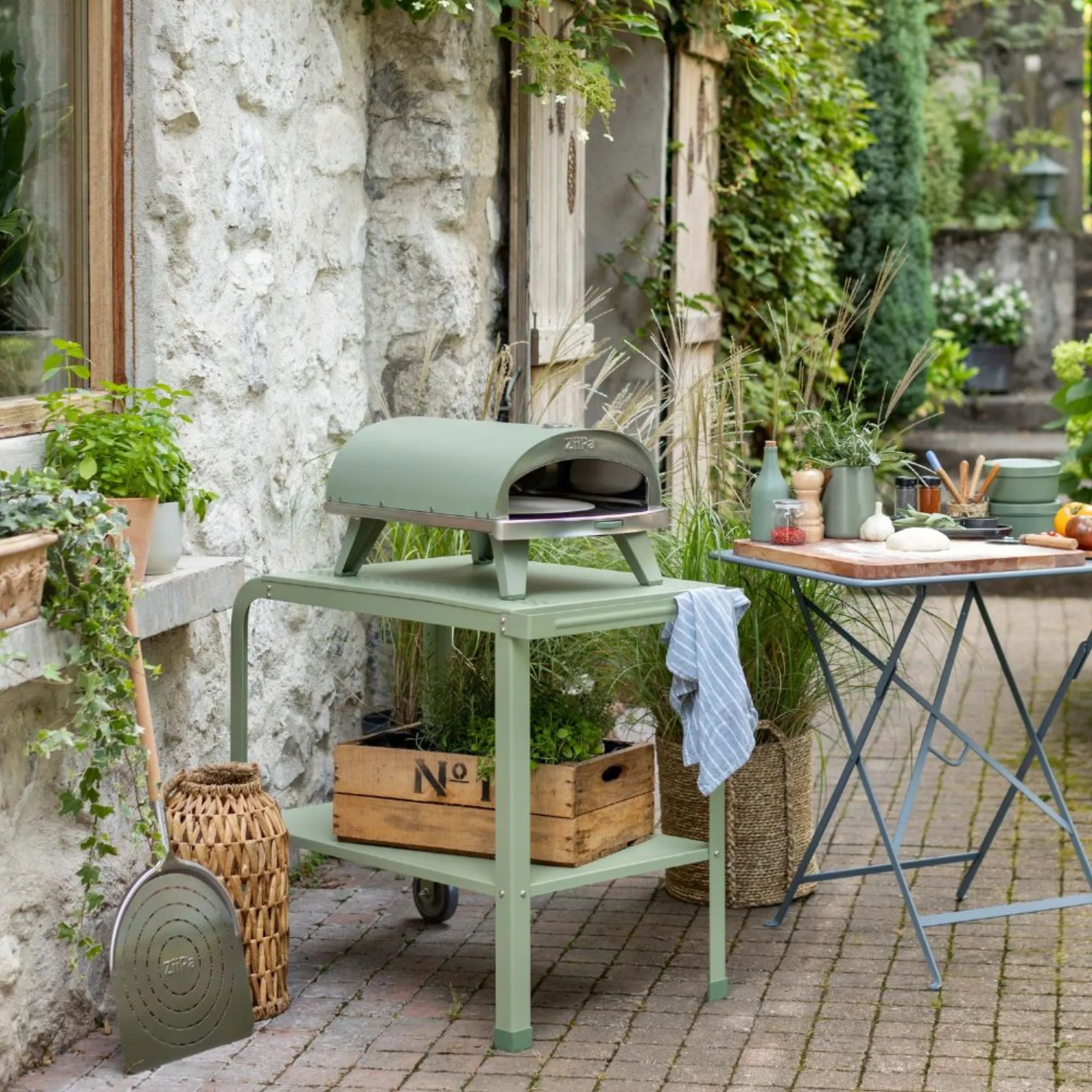 ZiiPa Piana wood-fired pizza oven on a vallone trolley, both in Eucalyptus colour, on a garden patio in front of a pizza-making station
