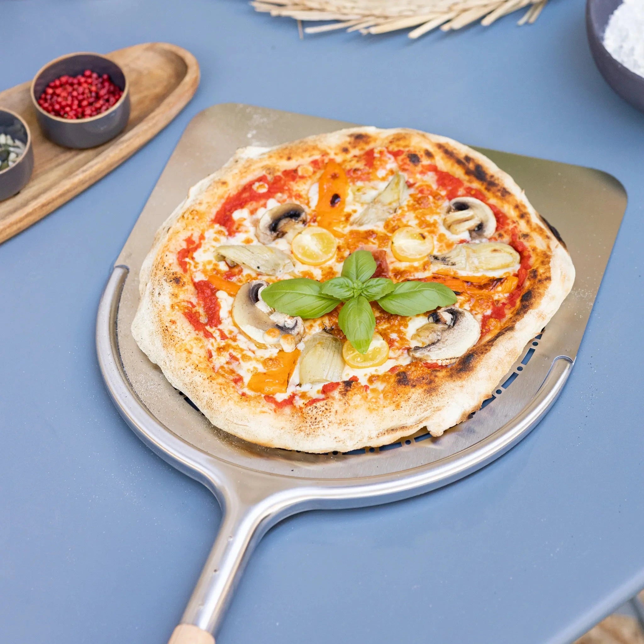 ZiiPa Aquila perforated pizza peel with a beech wood handle with a cooked pizza on a blue worktop