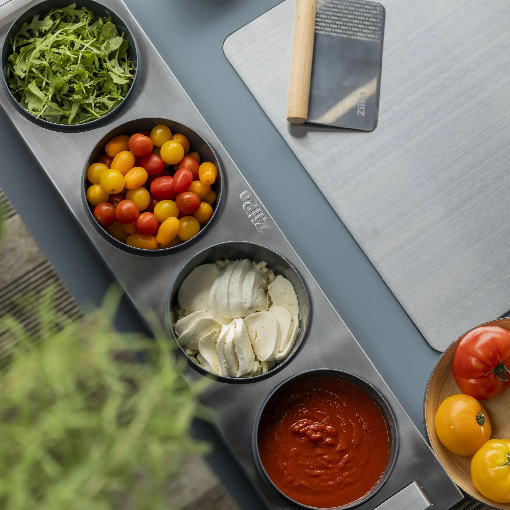 Top down view of a Tellini topping station with herbs, tomatoes, mozzarella and tomato sauce inside the charcoal spetina dough boxes