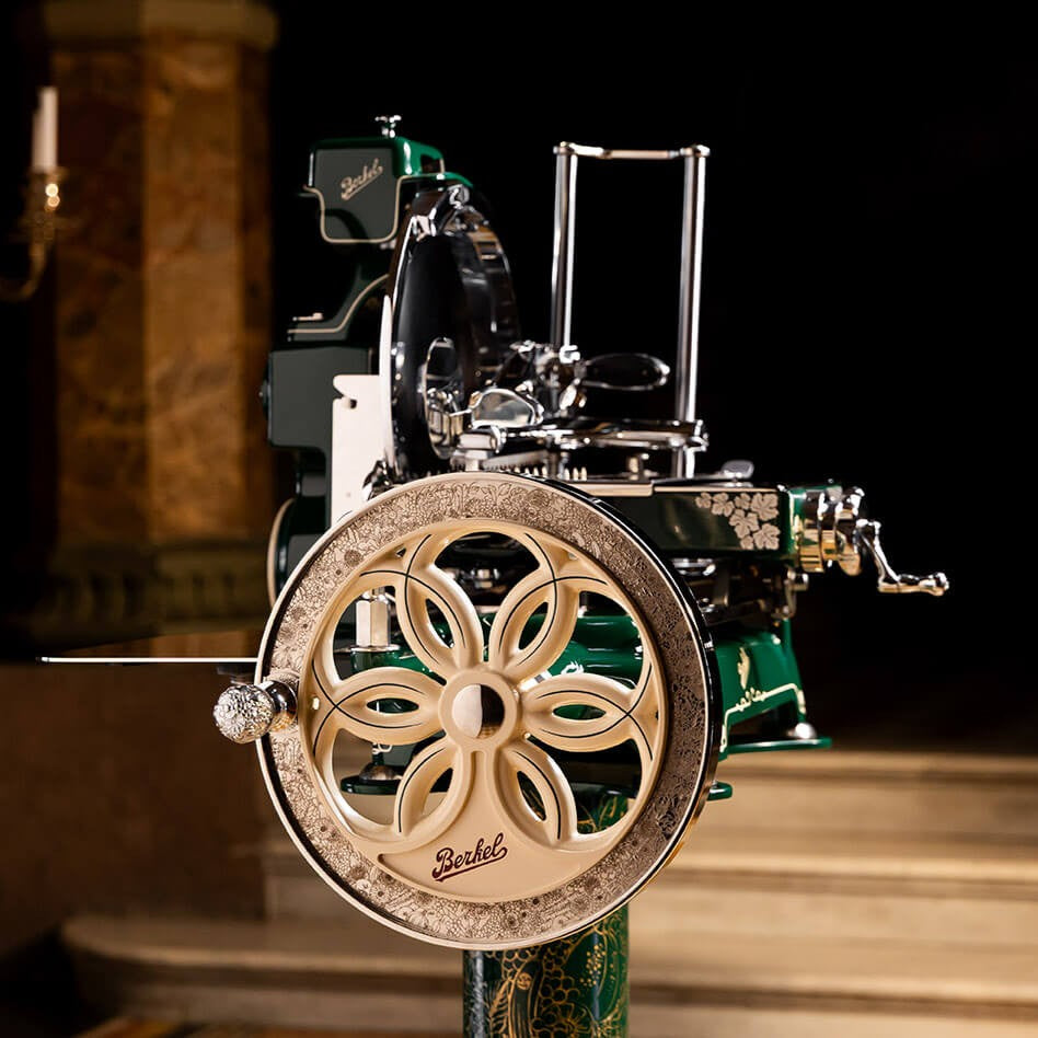 A close-up of a decorative vintage-style flywheel slicer with a large ornate beige wheel, polished silver metal parts, and green accents, photographed against a dark indoor background with blurred architectural elements.
