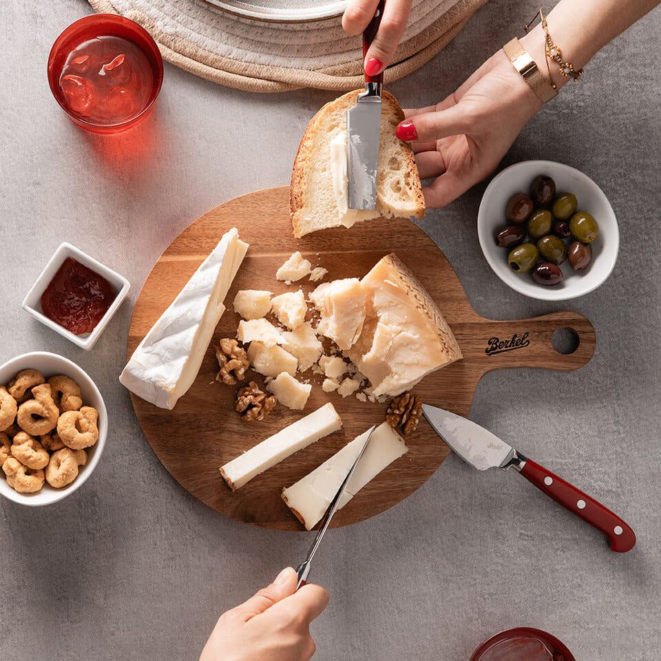 Person spreading cheese on bread with a Berkel knife on a Berkel board with various cheese and snacks on a gray surface