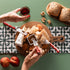 Hands preparing food on a Berkel branded cutting board with bread, fruits, and a jar of jam on a green surface.
