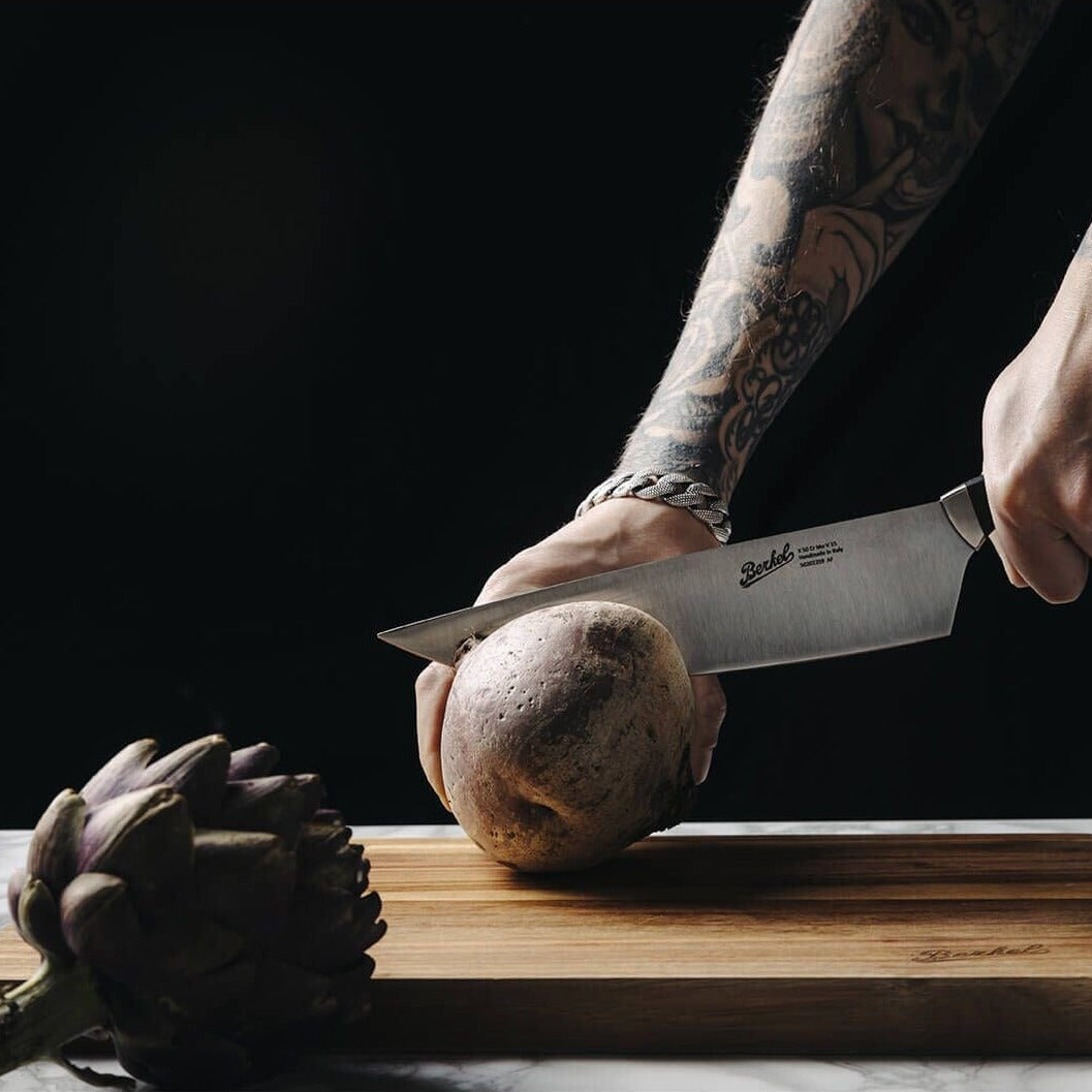 Person cutting an artichoke on a Berkel acacia chopping board with a black Berkel Teknika knife, against a black background