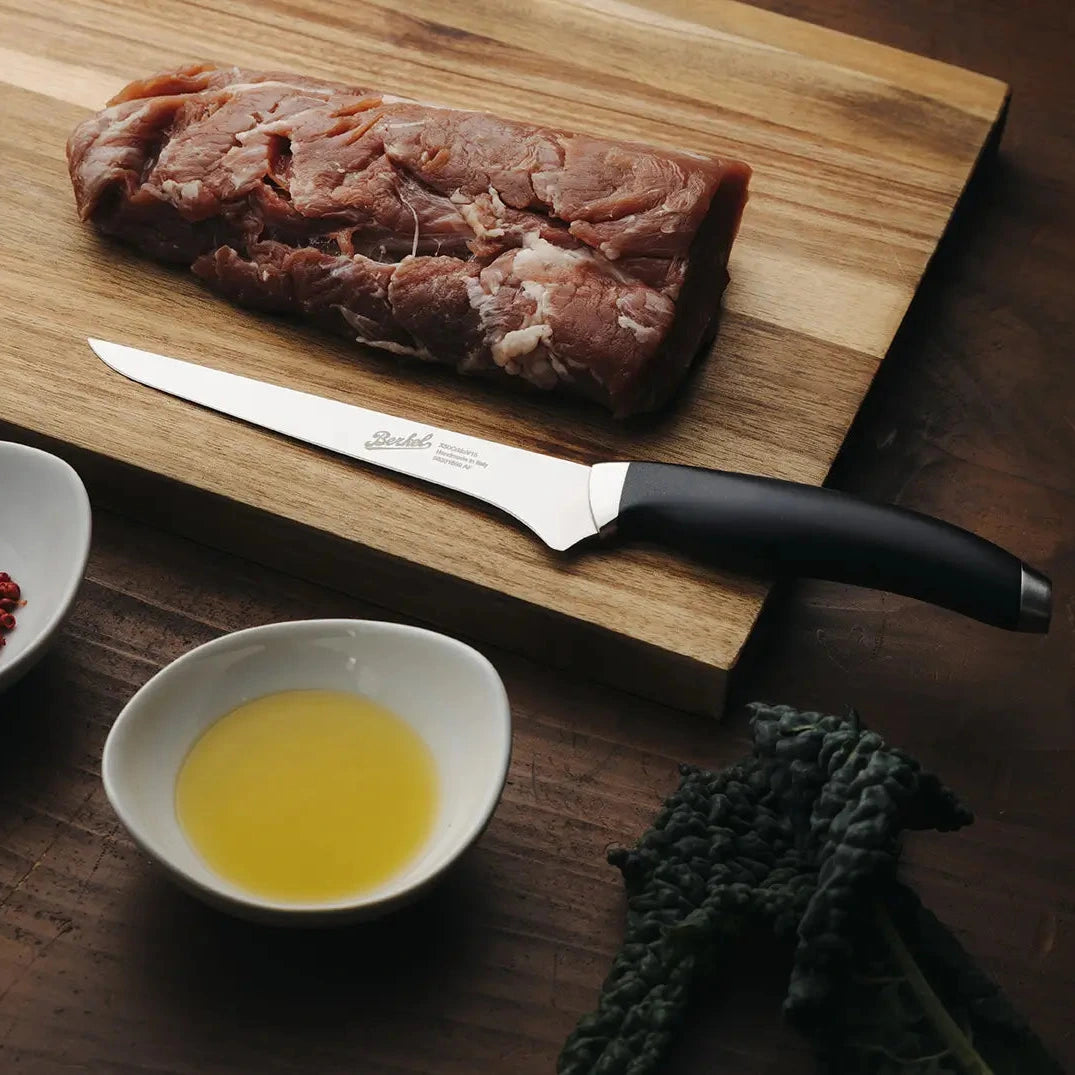 Steak on a wooden Berkel cutting board with a knife, bowls of spices and oil, and a leafy vegetable on a wooden surface.