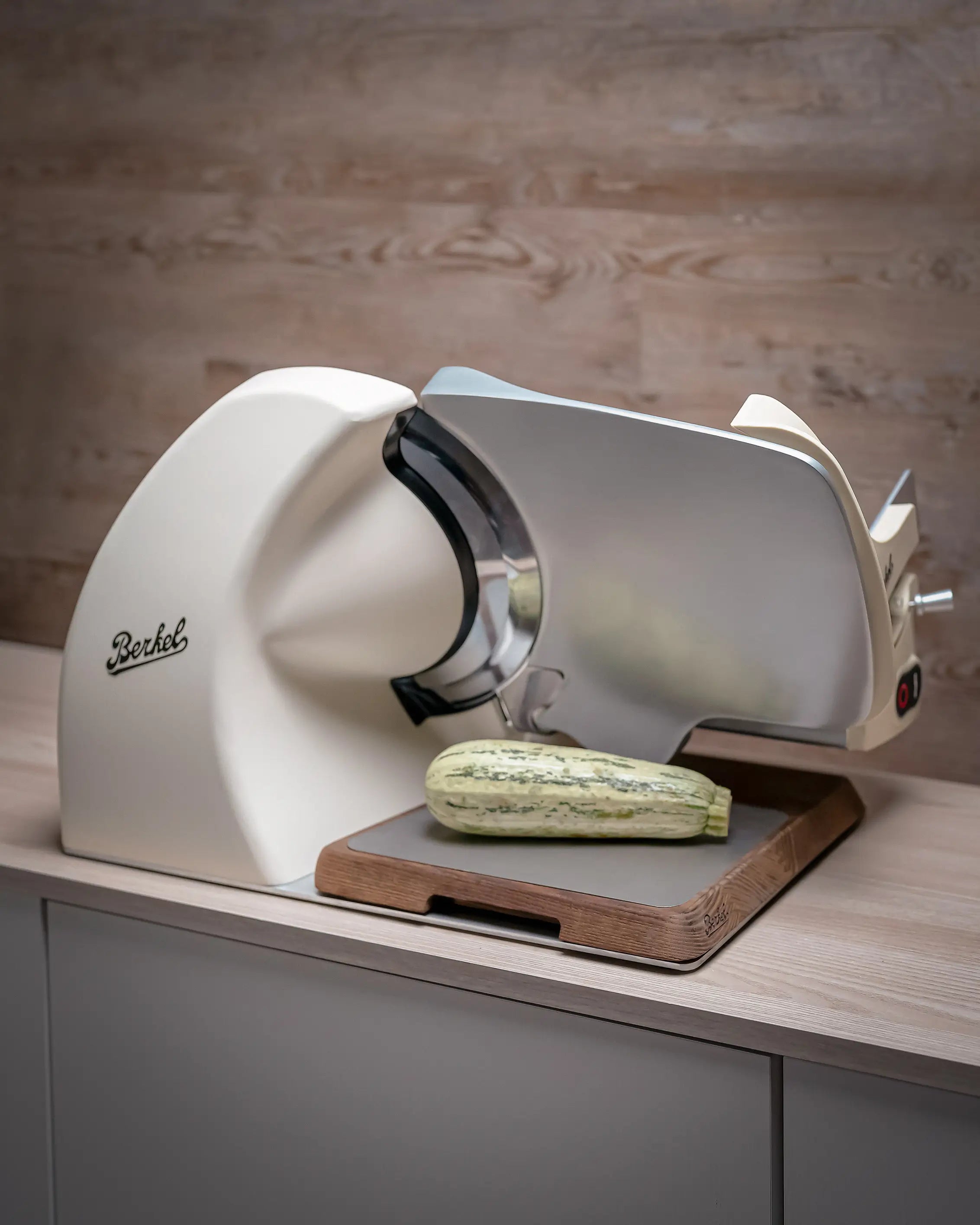A cream-coloured Berkel electric meat slicer shown in a kitchen setting, slicing a courgette on a wooden board.