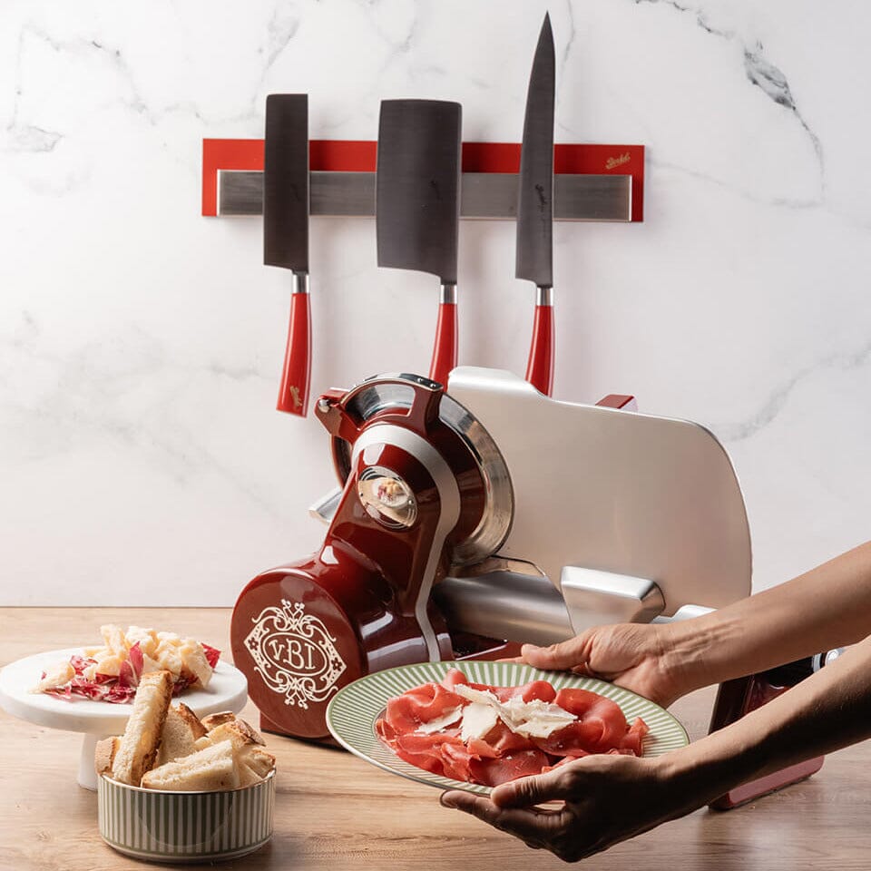 Person using a Berkel Icon line slicer on a wooden worktop with a magnetic knife holder with Berkel elegance knives on a marble wall.